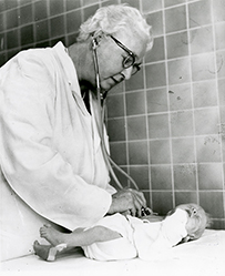 Dr. Virginia Apgar, a White female in a lab coat and glasses using a stethoscope to listen to a infant’s heart.