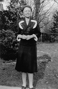 Dr. Emily Partridge Bacon, a White female in a blouse and skirt standing outdoors near shrubs.