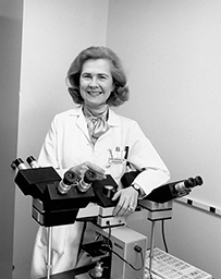 Dr. Doris Gorka Bartuska, a White female in a lab coat posing with a four-headed microscope.