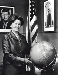 Dr. Leona Baumgartner, a female posing in a room with a globe, JFK's portrait, and the American flag.