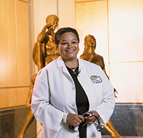 Dr. JudyAnn Bigby, an African American female in a lab coat holding a stethoscope in front of a sculpture.