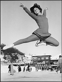 Dr. Lorraine Hanlon Comanor, a White female figure skater posing in a figure-four jump in the air with her arms overhead.