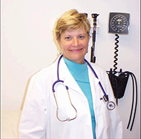 Dr. Marie Amos Dobyns, a White female in a lab coat with a stethoscope around her neck standing in front of a blood pressure measure.