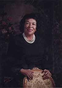 Dr. Roselyn Payne Epps, a smiling African American female wearing a triple strand of pearls posing next to a chair for her portrait.