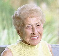 Dr. Jane E. Hodgson, an elderly, smiling White female seated at a blue table holding a newspaper.