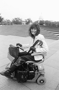 Dr. Lisa I. Iezzoni, a White female with curly hair seated on an electric scooter outdoors in front of steps. The Capitol building in D.C. is in the background. 