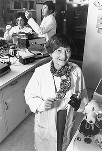 Dr. Lissy Feingold Jarvik, a White female wearing a lab coat next to a microscope in her laboratory with Nalini Makhijani and Steve Matsuyama in the background.