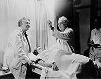 Dr. Catherine Macfarlane, a White female in a lab coat to the left of a patient on a bed and a two males on the right.