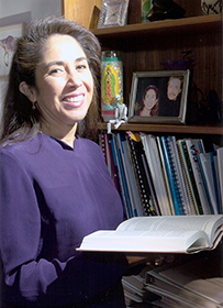 Dr. Martha Alicia Medrano, a Latina female holding a book open while standing in front of bookshelves.