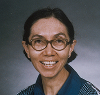 Dr. Fernande Marie Pelletier, a smiling female wearing a blue patterned dress and glasses for her portrait.