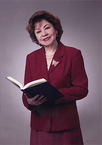 Dr. Sylvia M. Ramos, a smiling female in a red suit and skirt, holding an open book, posing for her portrait.