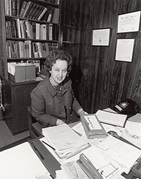 Dr. Marianne Schuelein, a White female posing at a desk with lots of papers and a bookshelf in the background.