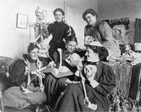 Dr. Ida Sophia Scudder, seated on left, holding skeleton arms bones, in a room with her female classmates, each posing with skeleton pieces.
