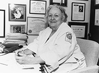Dr. Lucy Frank Squire, a White female seated at desk in a lab coat with medical degrees hanging on the walls.