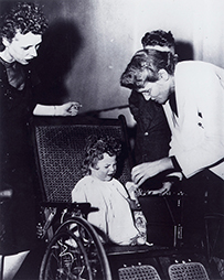 Dr. Helen Taussig, a White female leaning over to tend to a curly haired child seated in a wheelchair.