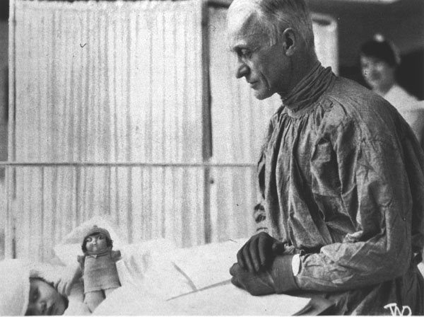 Black and white photograph of Dr. Harvey Cushing dressed in medical scrubs and wearing gloves standing at the bedside of a young patient lying on their side with bandages on their head and covered with white sheets. There is a baby doll leaning up against the patient.