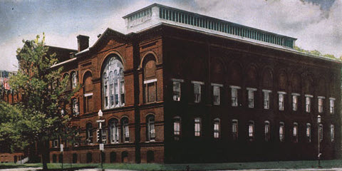 Exterior view of a red building. Trees line the front facade; U.S. flag hangs in front of the building; trolley tracks are in the foreground.
