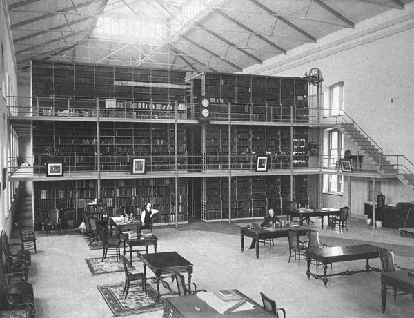 Interior view of Library and Museum of the Surgeon General's Office. John Shaw Billings is sitting at a table in the middle of Library Hall; Thomas W. Wise sits at a desk resting his elbow on the back of the chair. The three tier stacks are in the background, portraits are hanging off the second tier. A book truck with a duster is on the ground floor. Tables and chairs are in the foreground of the reading room. Windows run along the walls on all three tiers. There appears to be a sky light.