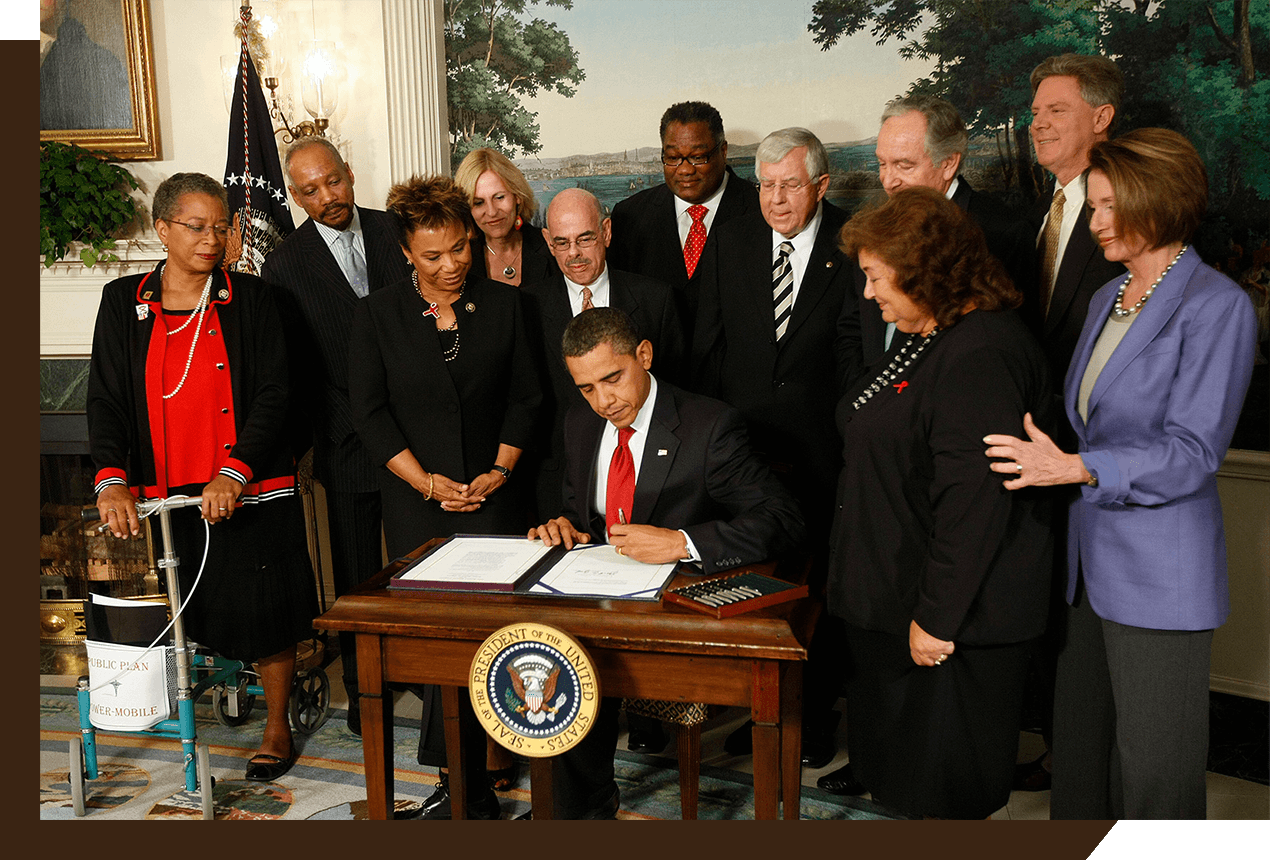 El Presidente Obama firmando un documento. Lo rodea un grupo de once personas.