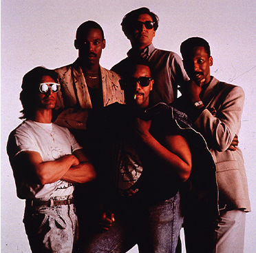 Grupo de cinco hombres, tres afroamericanos y dos blancos, todos mirando a la cámara; a la derecha se muestran dos condones.   