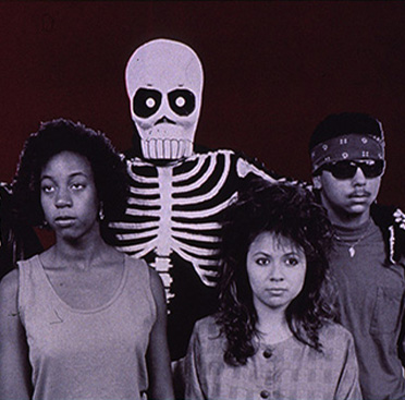 Un hombre y una mujer afroamericanos, un hombre y una mujer hispanos y, en medio de ellos y posando los brazos sobre los hombros, una persona en disfraz de esqueleto.