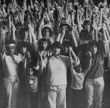 Black and white photograph with a multiracial group of adults and children holding each other’s hands above their heads.  