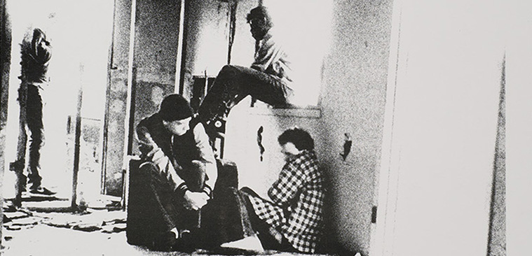 Black and white photograph of run-down front room of a house with three people huddled together, with a fourth person walking out the front door.