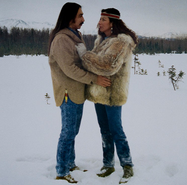 Cartel con texto y la fotografía a color de un hombre y una mujer nativos de Alaska abrazados en un campo cubierto de nieve.