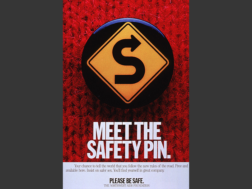 Color photograph of a safety pin on a red sweater; the pin has a yellow roadside on it