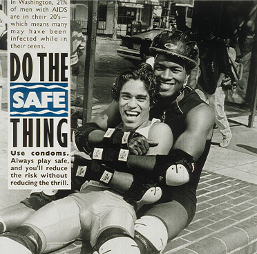 African American man sitting down and embracing a Latino man; both are wearing knee and elbow pads and one is wearing a helmet and rollerblades