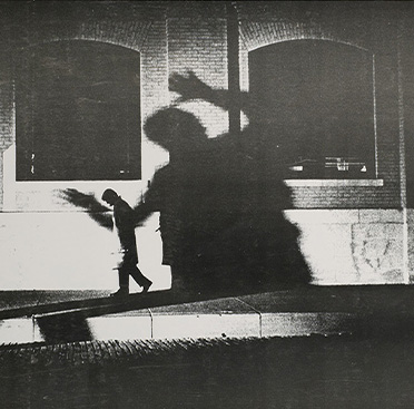 Black and white photograph of man walking at night, shadow behind him on building is exaggeratedly large and portrays a man about to grab someone