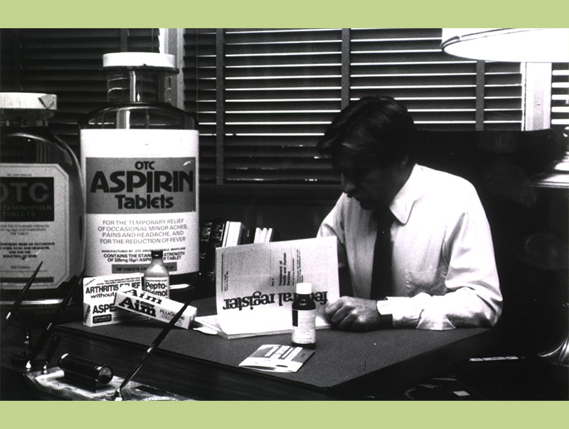 A black and white photograph of a white man reading a document while sitting at a desk with containers of varying size of over the counter medicines