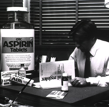 A black and white photograph of a white man reading a document while sitting at a desk with containers of varying size of over the counter medicines