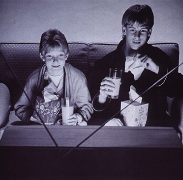 An advertisement showing text below a black and white photo of two white, adolescent boys sitting on a couch in front of a television and holding tissue boxes and glasses of orange juice