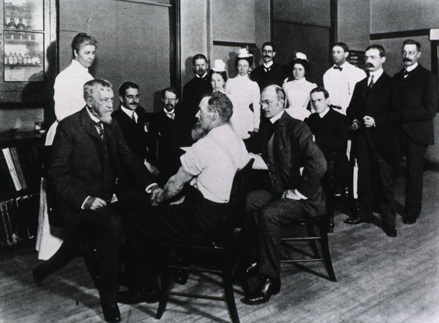 Dr. Mitchell examining a male patient in left foreground while a group of men and women look on.
