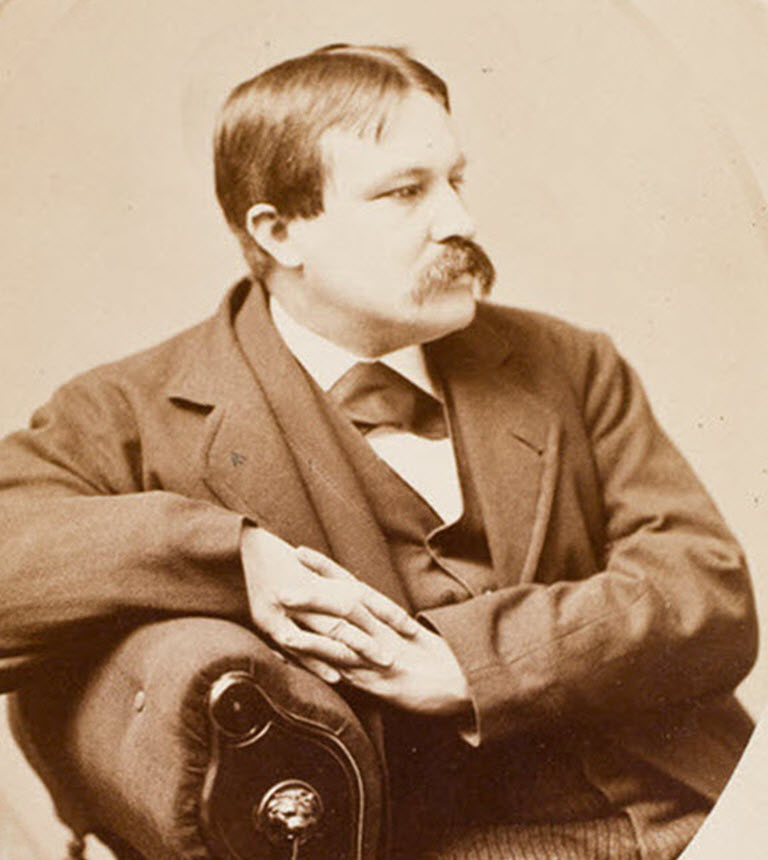 A man with a mustache wearing suit and leaning on a chair arm in profile facing the right.