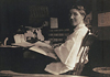A woman at seated at a desk, turning towards her left to look at viewer.