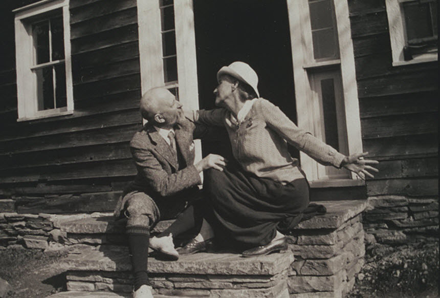 An older man and woman smiling at each other and sitting on stairs in front of a building.