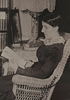 A woman reading in a wicker chair, next to shelves of books.