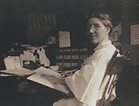 A woman at seated at a desk, turning towards her left to look at viewer.