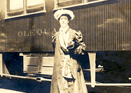 A woman in dress and hat standing in front of a train car.