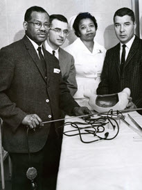 Three men in suits and a female nurse stand by a table with medical equipment.
