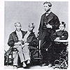 Photograph of Chang-Eng Bunker, seated, with two of their children, standing.  All are well-dressed in suits.