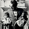 Dr. C. Everett Koop stands in a hospital room holding the separated Clara and Alta Rodriguez in each arm, prior to their discharge from the hospital. Both twins are wearing dresses and have bows in their hair.