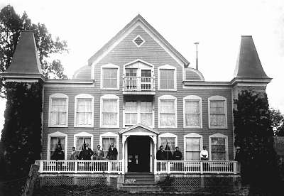 A black and white photo of Clara Barton's House, as it appeared in 1904, a multi-story wood home with a large front porch, 16 windows on the front with a wide stairs leading up either side to a six Greek ionic columned entrance.