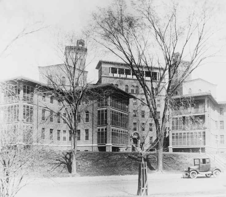 A black and white front angle view of the Columbia Hospital for Women, ca. 1920s - several multi-story buildings surrounded by trees.  A very early model vehicle is parked on the street.