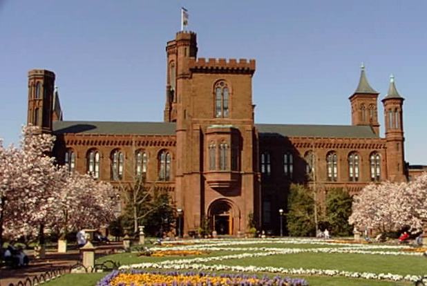 A color image of the front view of Smithsonian Institution - a multi-story brick building in the National Mall surrounded by Cherry Blossom trees and several colorful flower gardens on the grounds.