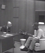 A black man in a baseball cap talks with a white man in a suit behind a desk in a wood paneled office.