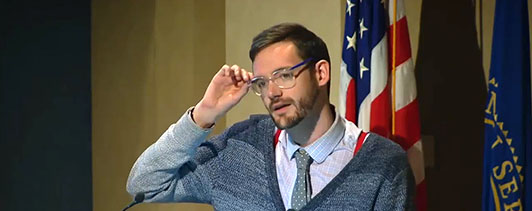 Young man at a podium adjusting his glasses.