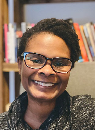 Photograph of a black woman in glasses in front of a bookshelf.